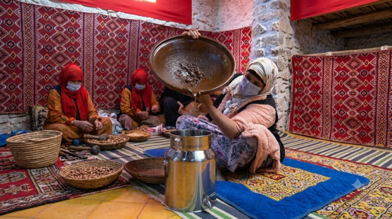 A candid photograph inside a traditional Moroccan Argan oil cooperative of Arganisme. Four women, wearing protective face masks and traditional clothing, are seated on patterned rugs working. In the foreground, a woman wears a light pink jacket and pours thick, crushed argan pulp from a large brown ceramic dish into a stainless steel milk container. Next to her are baskets filled with argan kernels. In the background, other women wear red headscarves and work with argan nuts against a wall decorated with traditional red and patterned textiles. The setting features stone walls and traditional decor.